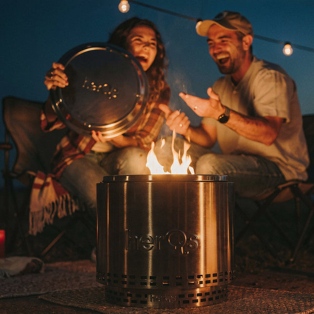 Lachend koppel buiten bij avond naast brandende herQs Cozy vuurkorf, vrouw houdt Closer deksel omhoog