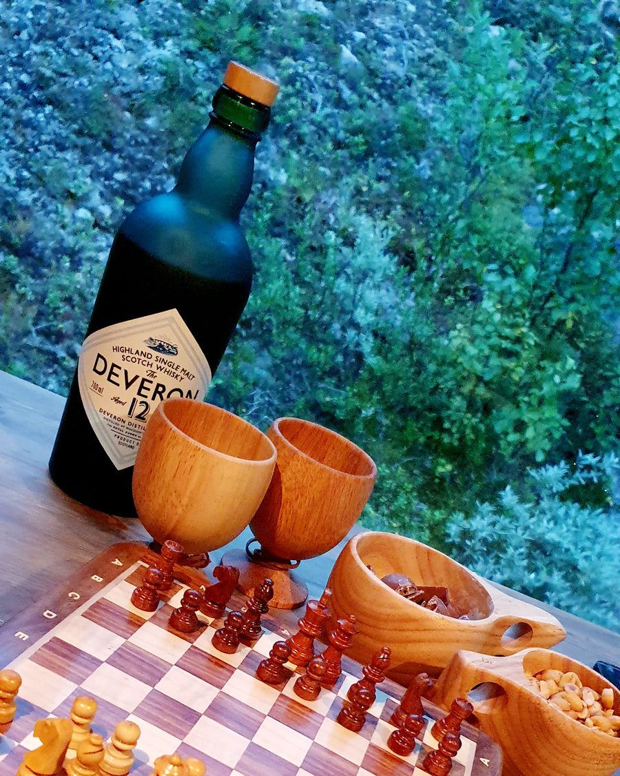 Twee Øyo houten wijnglazen naast een schaakbord en een fles buiten