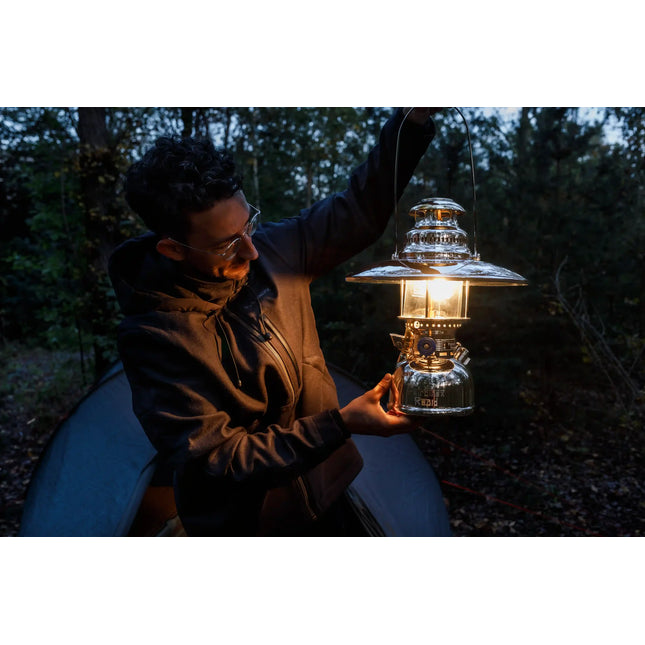 Man houdt een brandende Petromax HK500 met reflectorkap omhoog bij een tent in het bos